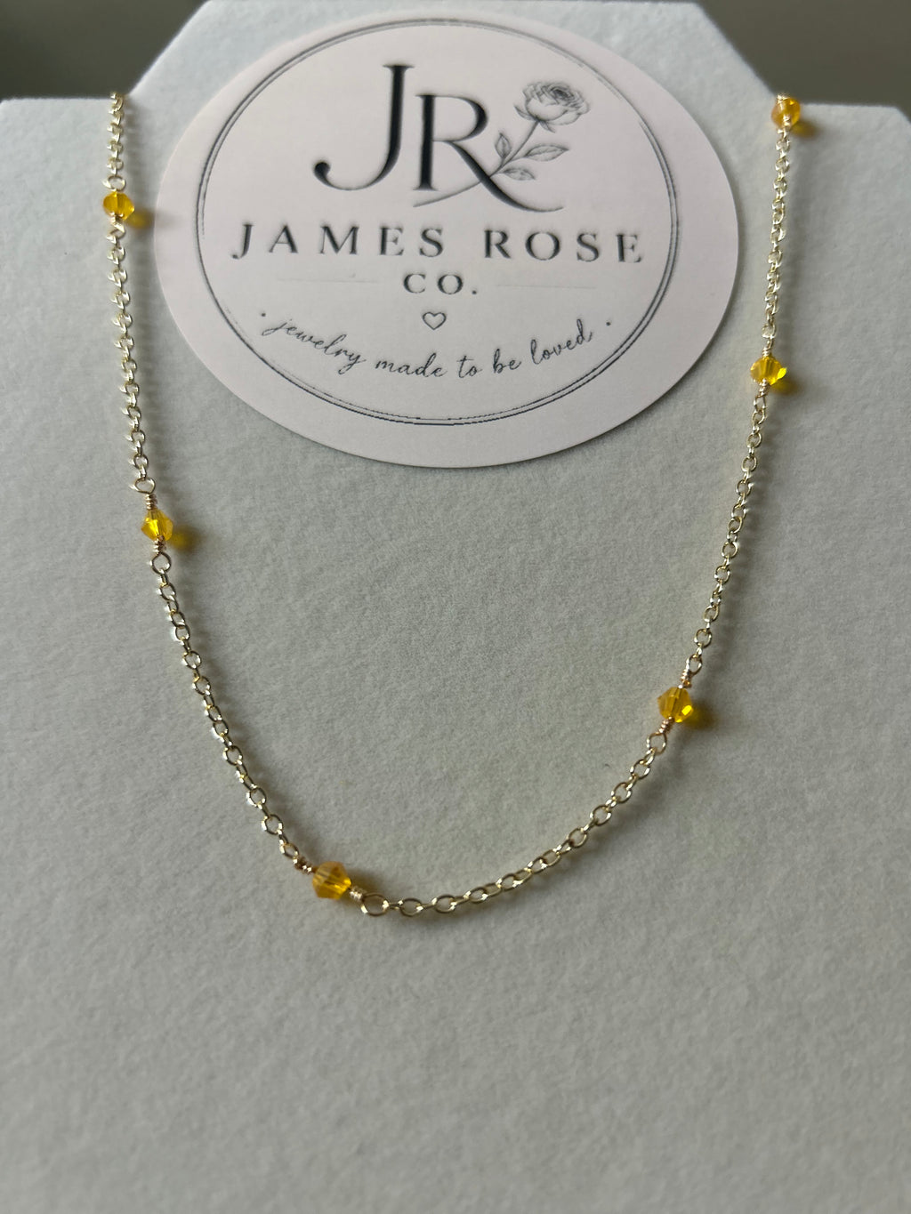 A delicate gold chain necklace with small yellow bead accents, displayed on a white jewelry stand with a circular James Rose Co. brand card behind it.