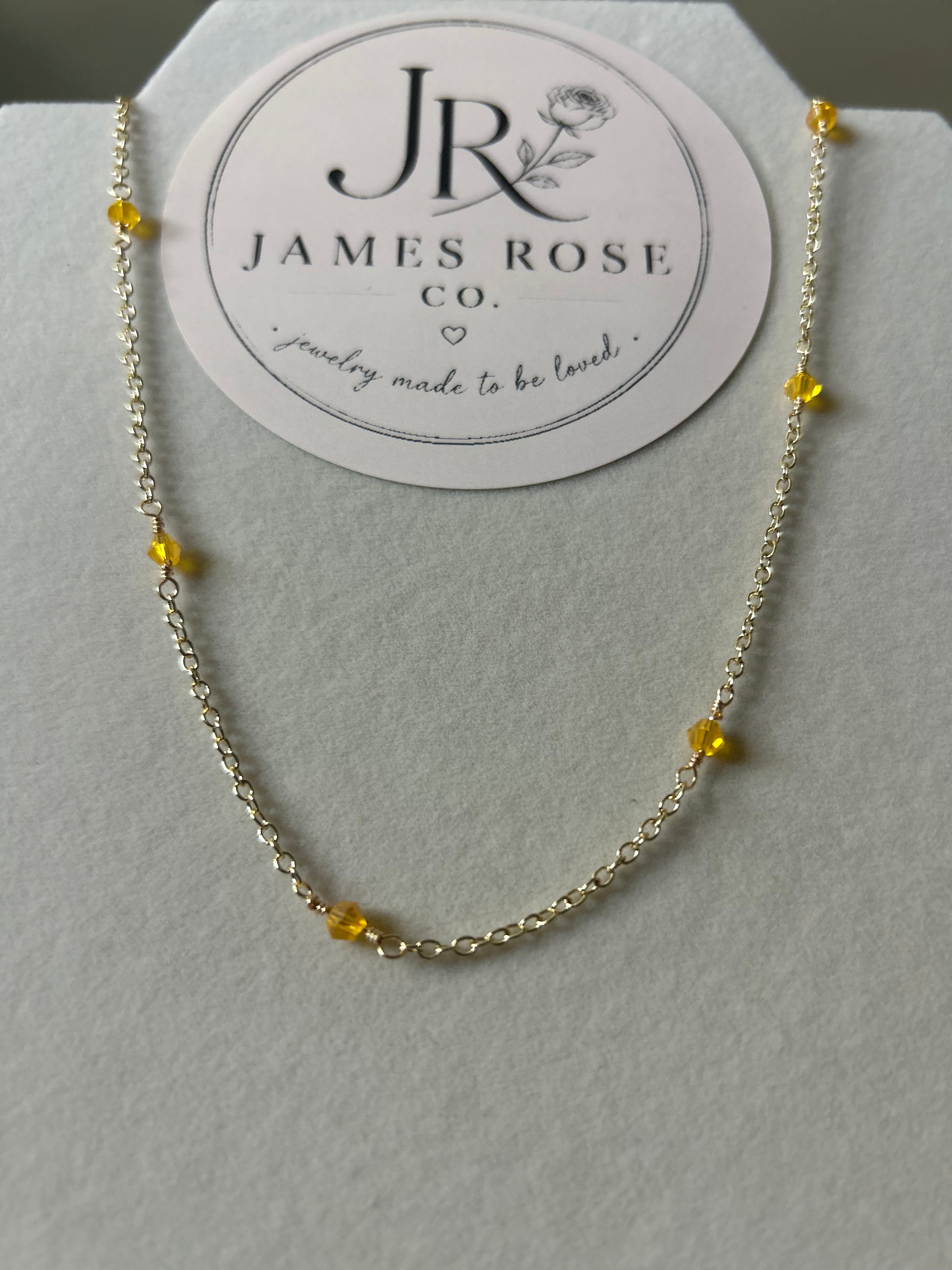 A delicate gold chain necklace with small yellow bead accents, displayed on a white jewelry stand with a circular James Rose Co. brand card behind it.