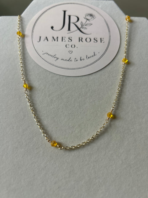 A delicate gold chain necklace with small yellow bead accents, displayed on a white jewelry stand with a circular James Rose Co. brand card behind it.